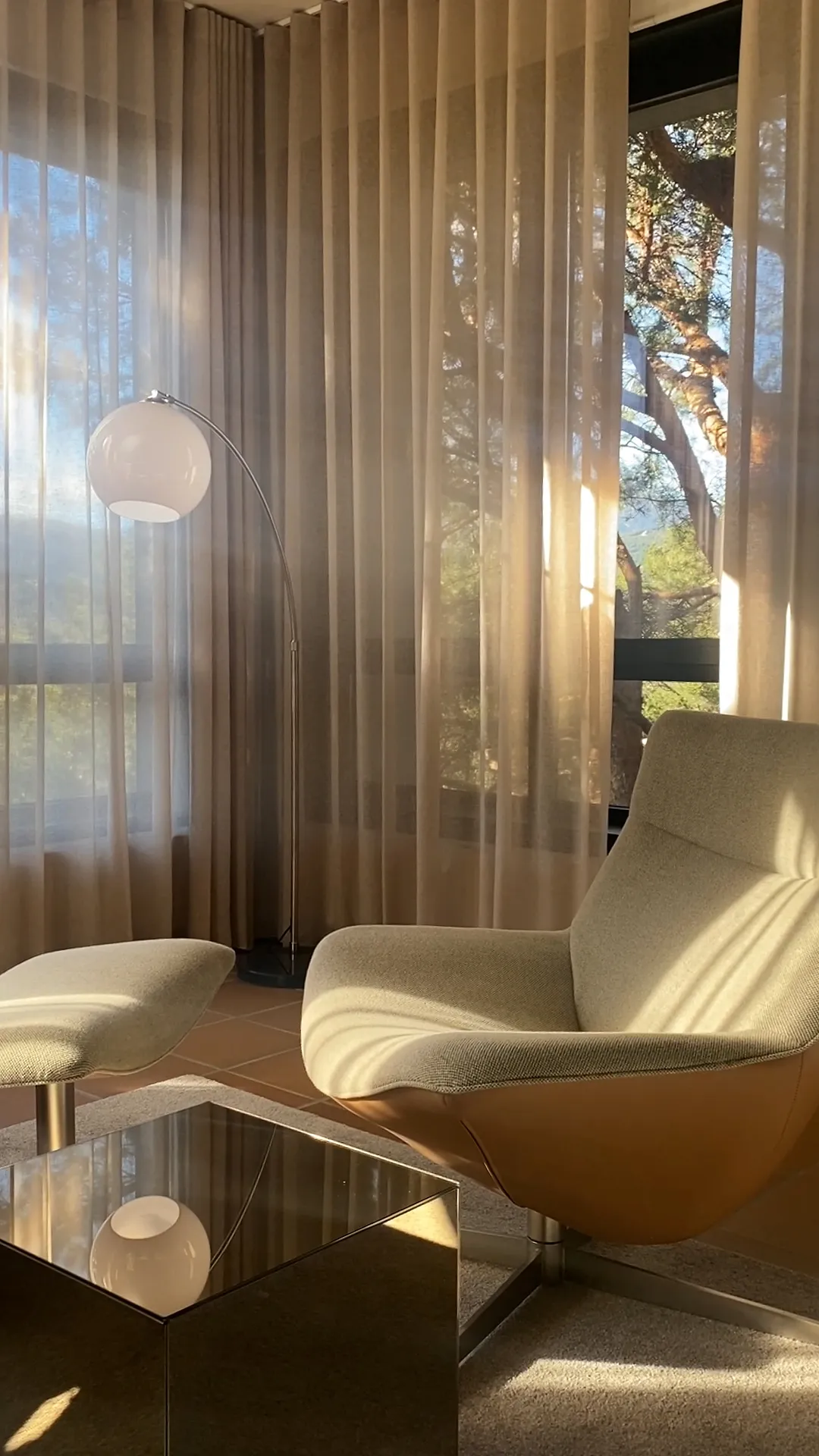 Modern living room corner with a beige armchair and matching ottoman next to a shiny cubic coffee table, a tall floor lamp with a round white shade, and sheer beige curtains covering windows showing trees outside in soft sunlight.
