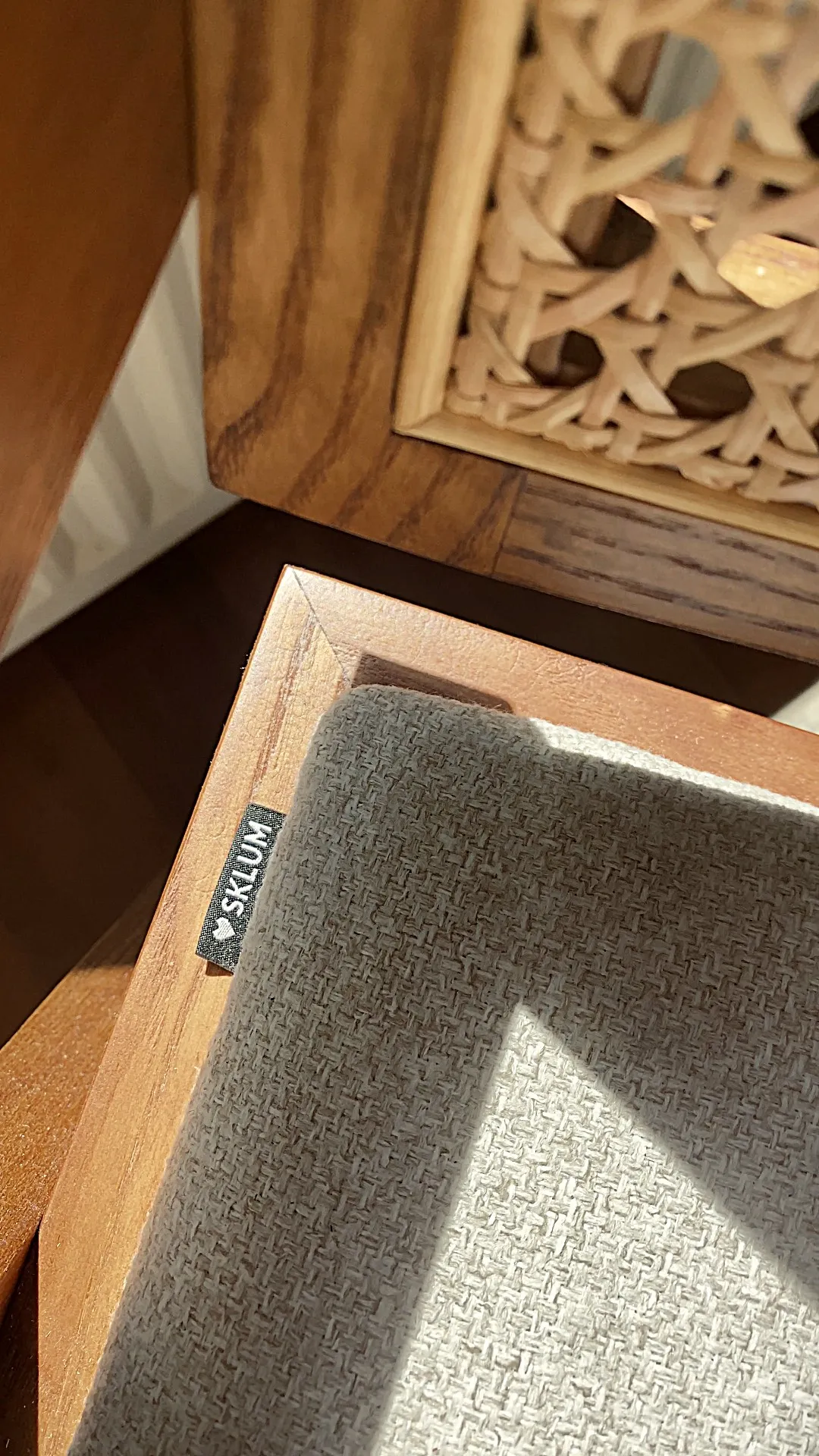 Close-up of a beige textured fabric cushion with a small black tag labeled 'SKLUM' resting on a wooden surface with another carved wooden object in the background.