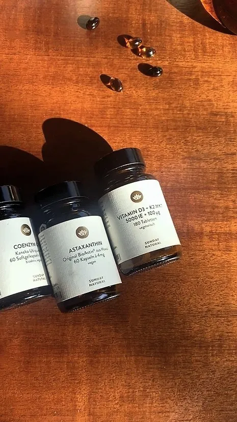 Three dark glass supplement bottles labeled Coenzyme Q10, Astaxanthin, and Vitamin D3 + K2 on a wooden surface with some softgel capsules scattered nearby.