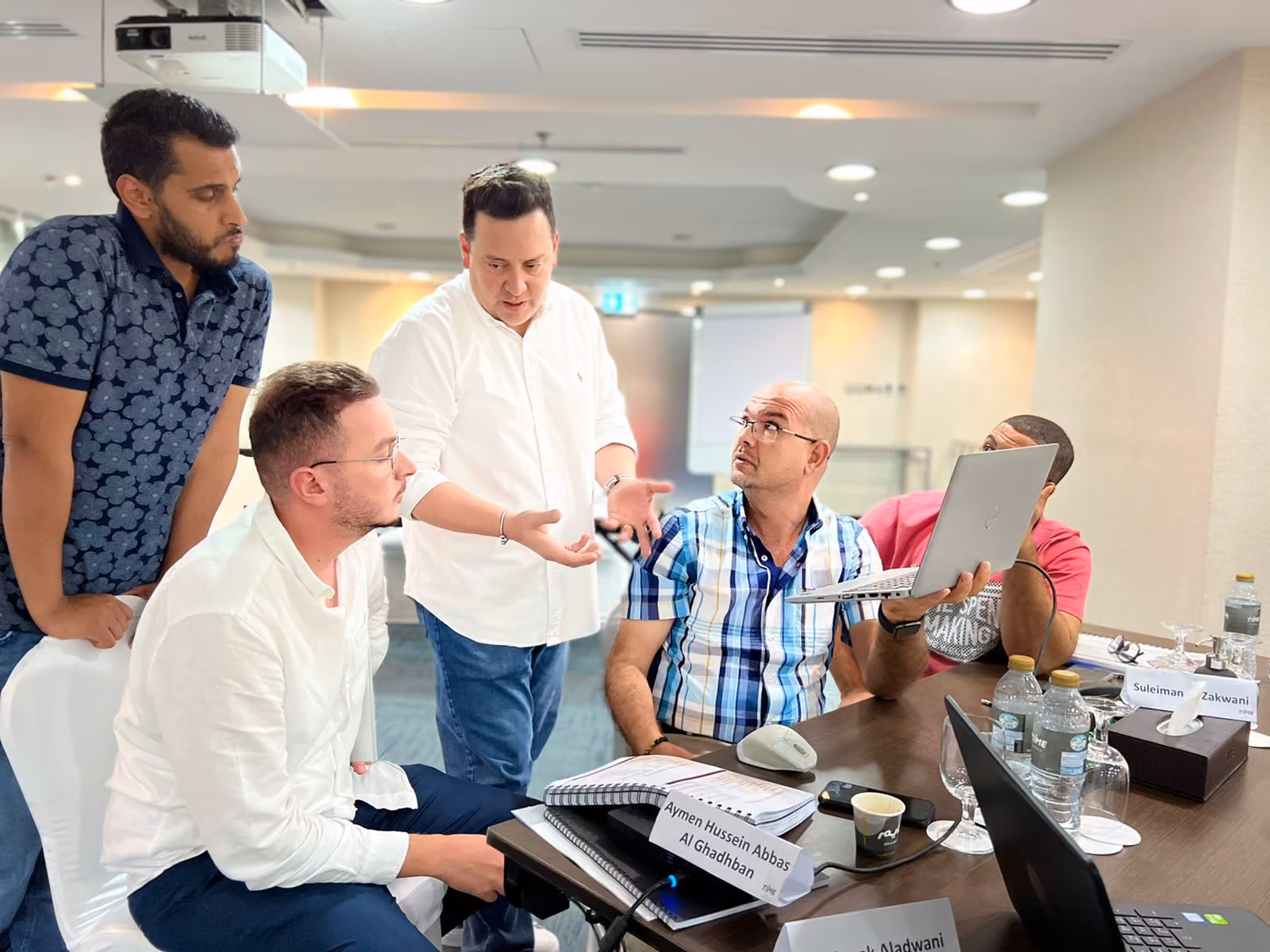 Five men engaged in a discussion around a table with laptops, notebooks, and bottled water.