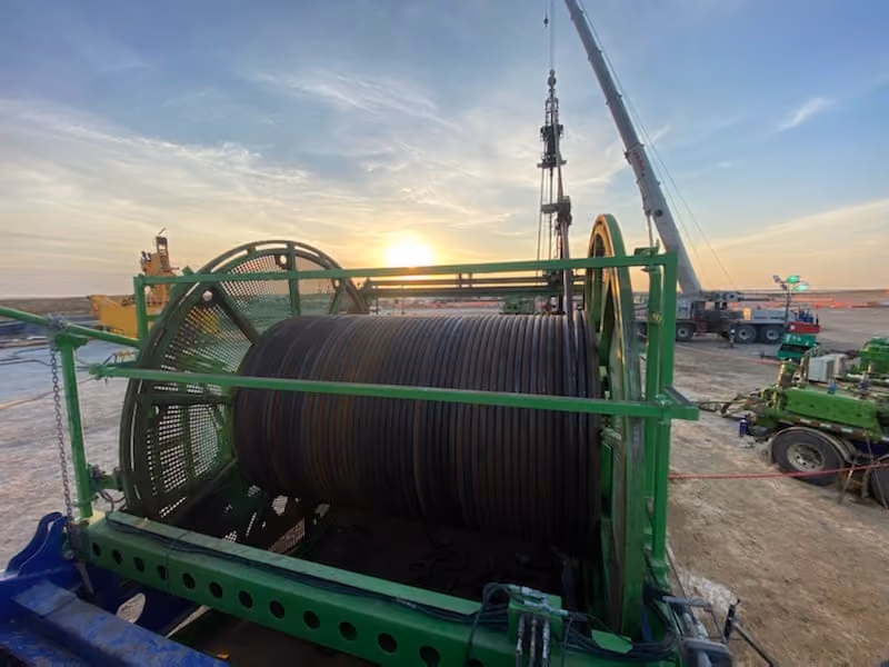 Large green industrial cable spool at a construction site during sunset with cranes and machinery nearby.