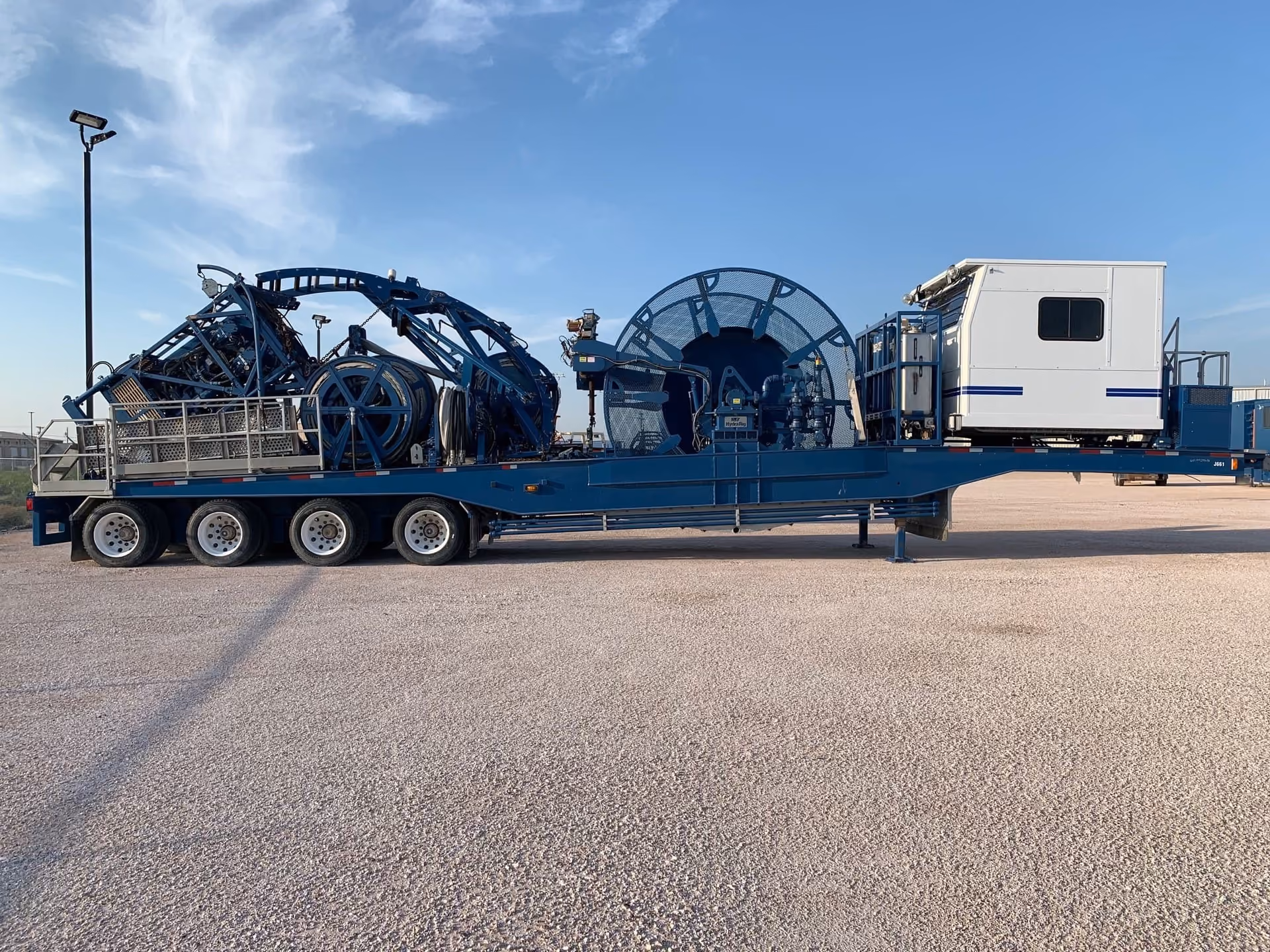 Large blue industrial trailer loaded with mechanical equipment and a white container unit under clear sky.