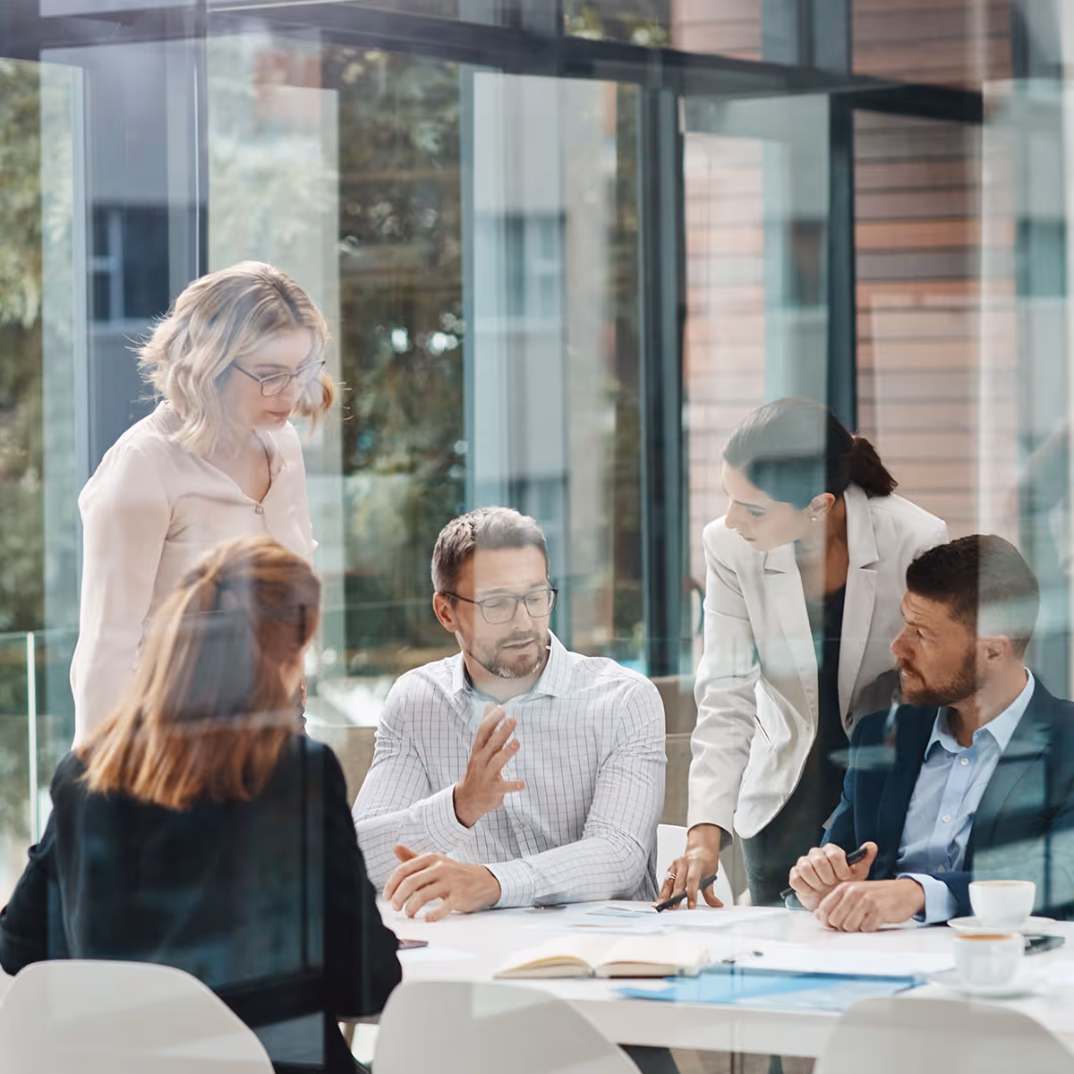 Five business professionals having a discussion around a white table in a modern office with large glass windows.