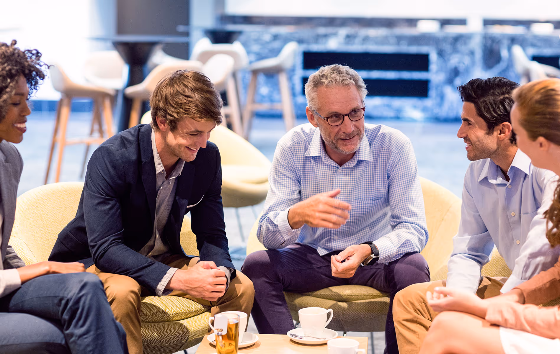 Five diverse professionals sitting in a casual office lounge, engaged in a lively group discussion with coffee cups on the table.