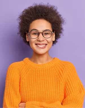 Smiling young woman with curly hair wearing glasses and an orange sweater against a purple background.