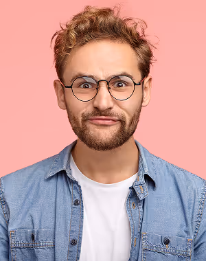 Young man with curly hair, glasses, beard, and denim shirt making a skeptical facial expression against a pink background.