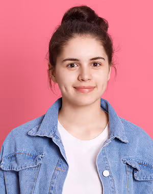 Young woman with dark hair tied in a bun, wearing a denim jacket and white shirt, smiling against a pink background.