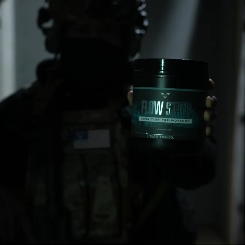 Person in tactical gear holding a black container labeled 'Flow State Cognition Pre-Workout' by Team Two Co.