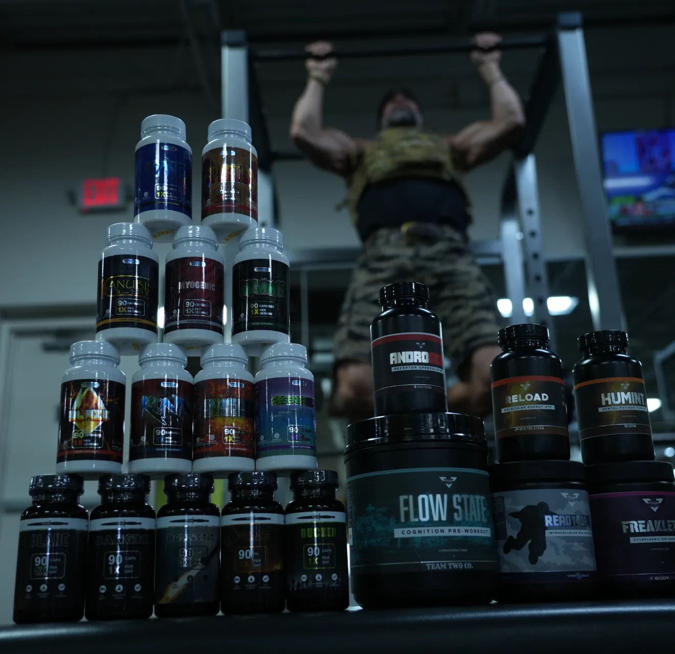 Stacked bottles of dietary supplements and nutrition products in a gym, with a man in camouflage workout clothes doing pull-ups in the background.