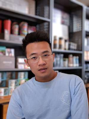 Young man with glasses wearing a light blue shirt, sitting indoors with shelves of various products in the background.