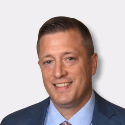 Smiling man with short brown hair wearing a blue suit jacket, light blue shirt, and patterned tie against a light background.