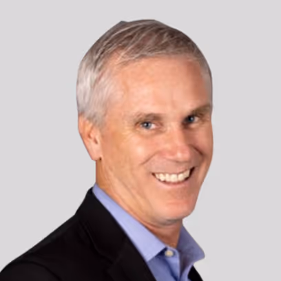 Smiling middle-aged man with short gray hair wearing a black blazer and blue shirt against a light gray background.
