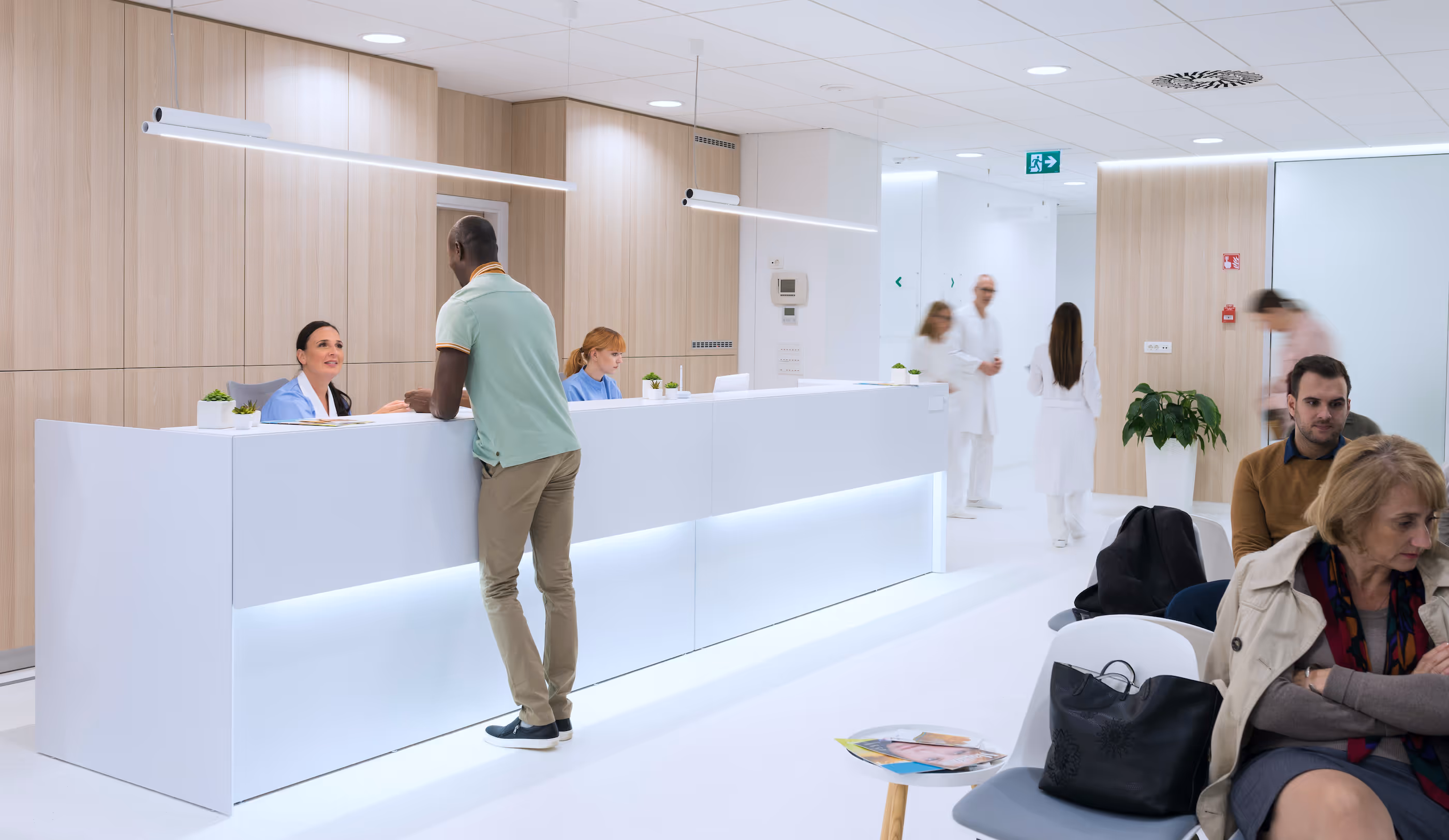 Modern medical office lobby with a man talking to a receptionist, patients waiting, and medical staff walking in the background.