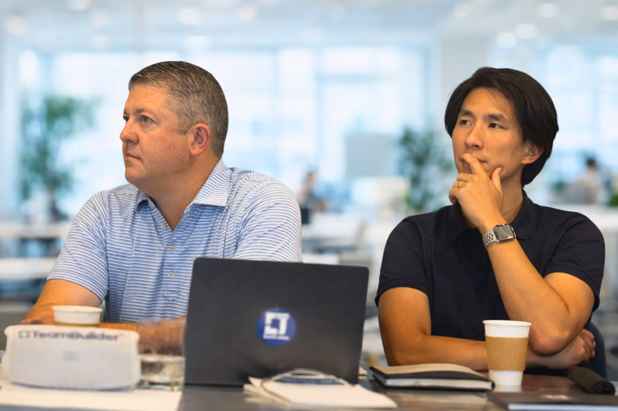 Two men sitting at a table in an office, one wearing a blue striped shirt looking left, the other in a black shirt with hand on chin looking thoughtful.
