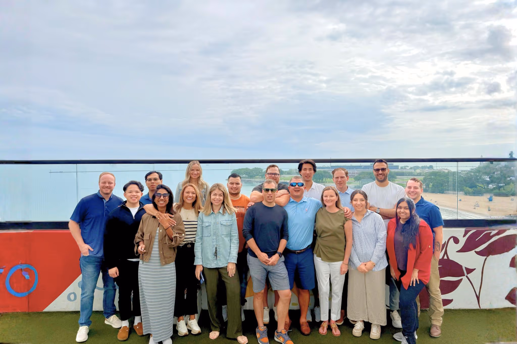 Group of 18 diverse adults standing and smiling on a rooftop with a cloudy sky and water in the background.
