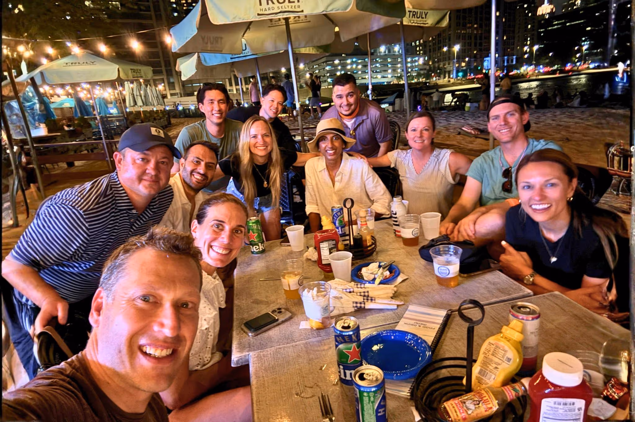 Group of eleven people smiling and seated around a table with drinks and condiments at an outdoor nighttime gathering.