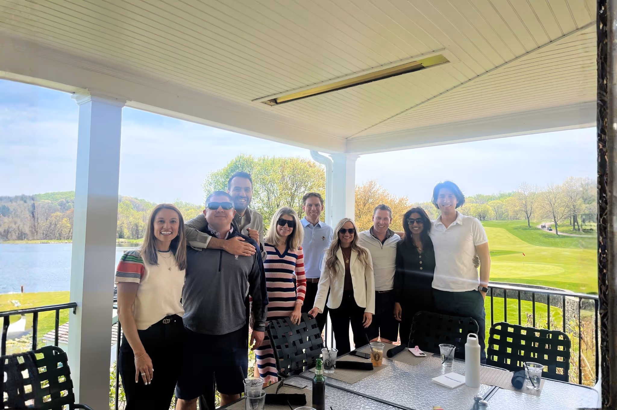 Group of nine people smiling and posing for a photo on a covered outdoor patio overlooking a golf course and lake.
