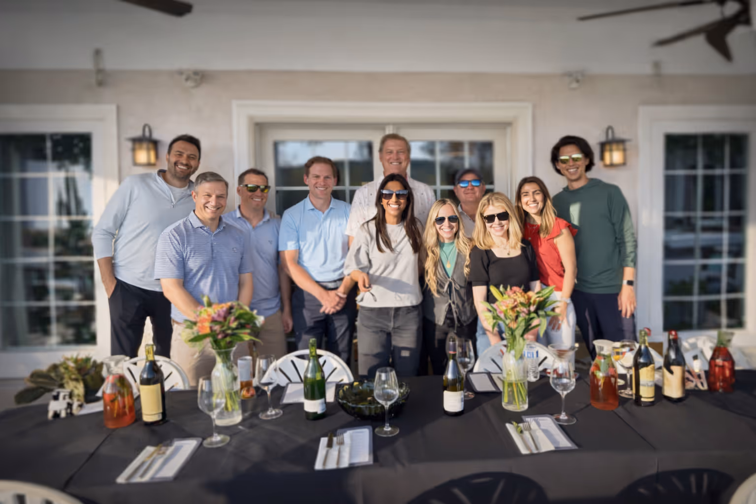 Group of smiling people wearing sunglasses standing behind a table set with flowers, wine bottles, and glasses outdoors.