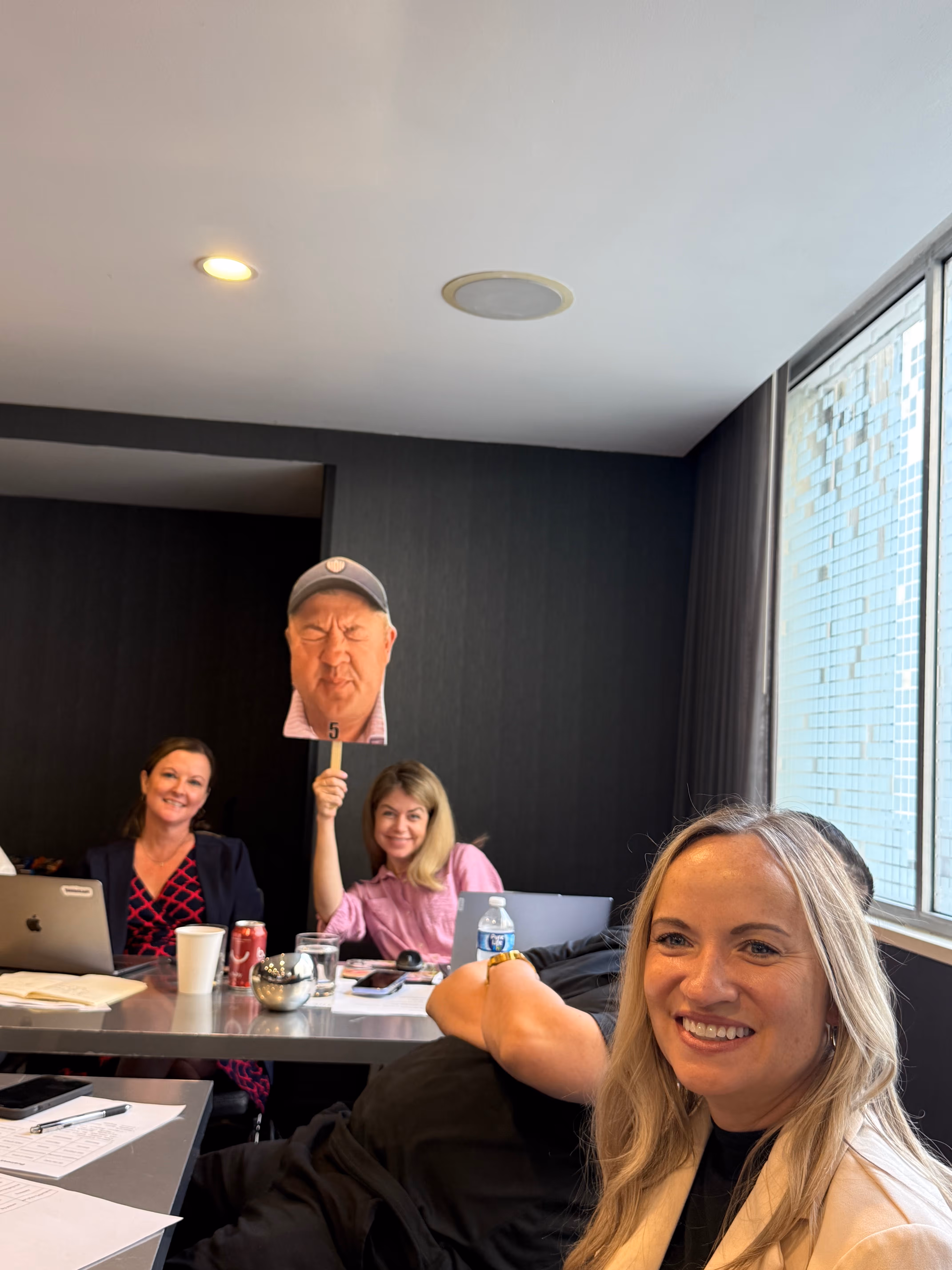 Four people sitting in a meeting room, one woman smiling in the foreground and another person holding a cutout of a man's face on a stick.