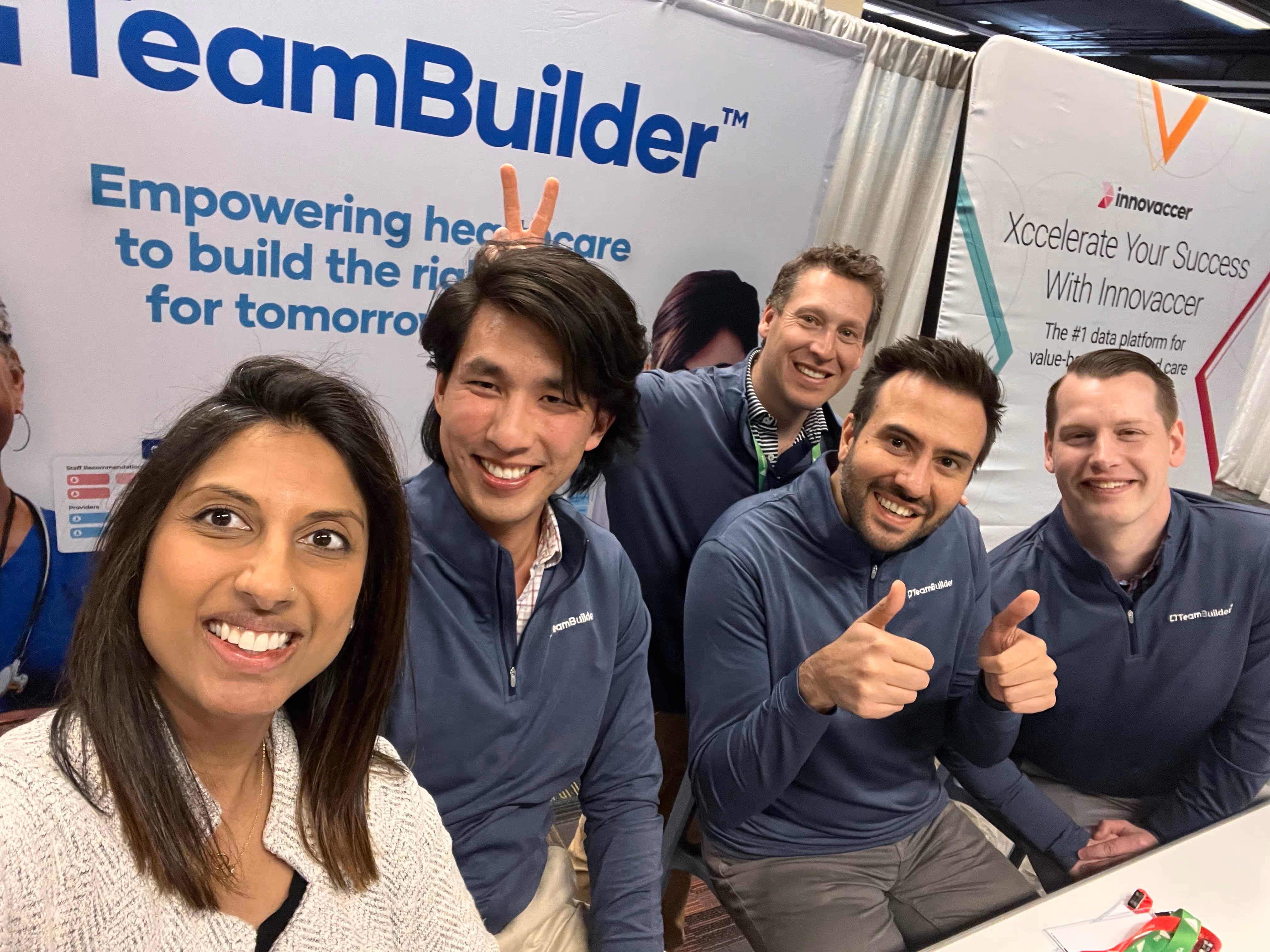 Five smiling people posing for a group photo at a TeamBuilder booth with banners promoting healthcare empowerment and Innovaccer.