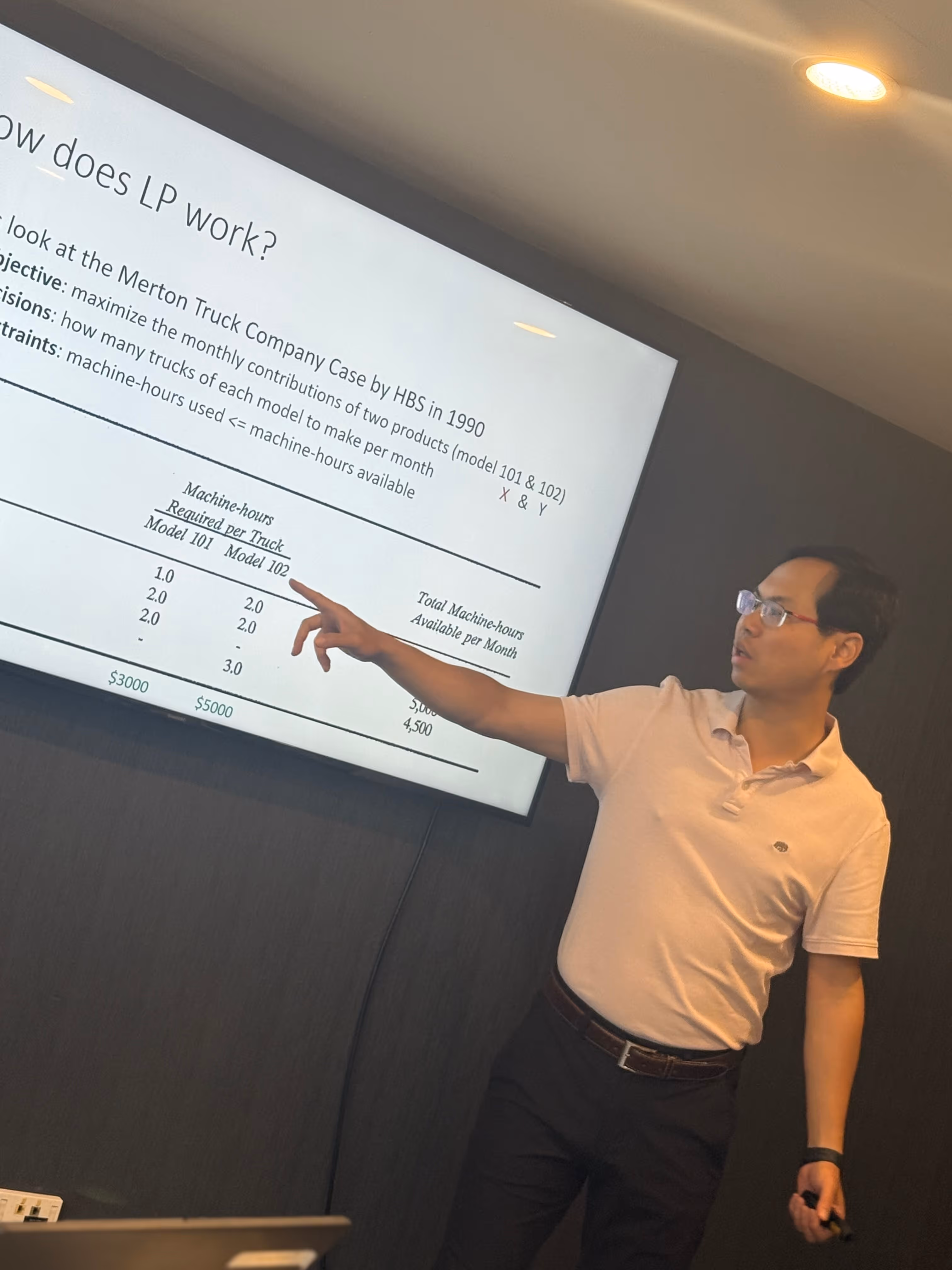 Man in glasses and white polo shirt pointing at a presentation slide about a Merton Truck Company case study on linear programming.