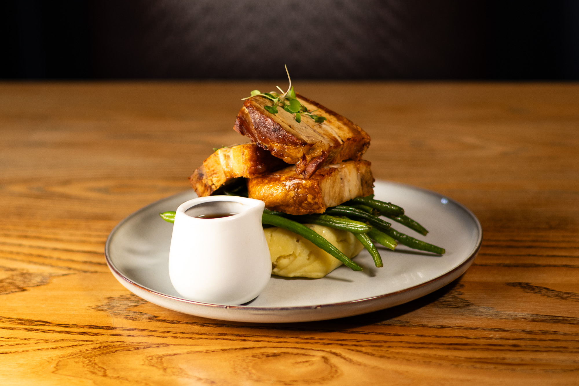 Plate with roasted pork belly slices garnished with microgreens, served on green beans and mashed potatoes with a small white sauce pitcher.