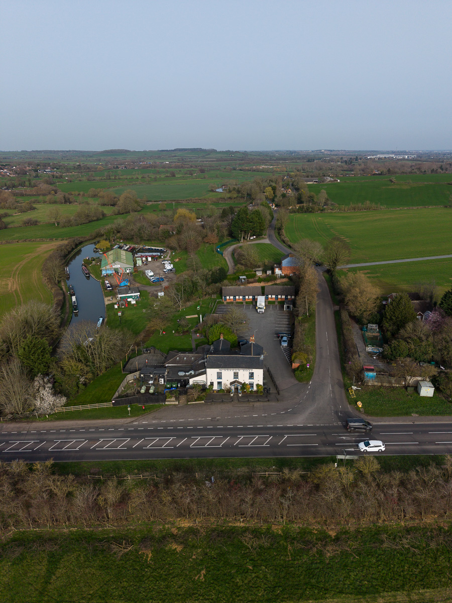 Aerial view of a rural pub beside a parking lot and narrow canal with boats, surrounded by green fields and trees.