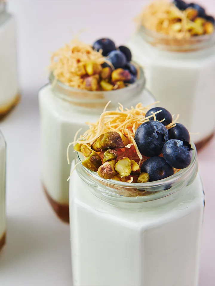 Individual dessert jars of creamy panna cotta topped with blueberries, chopped pistachios and golden shredded pastry