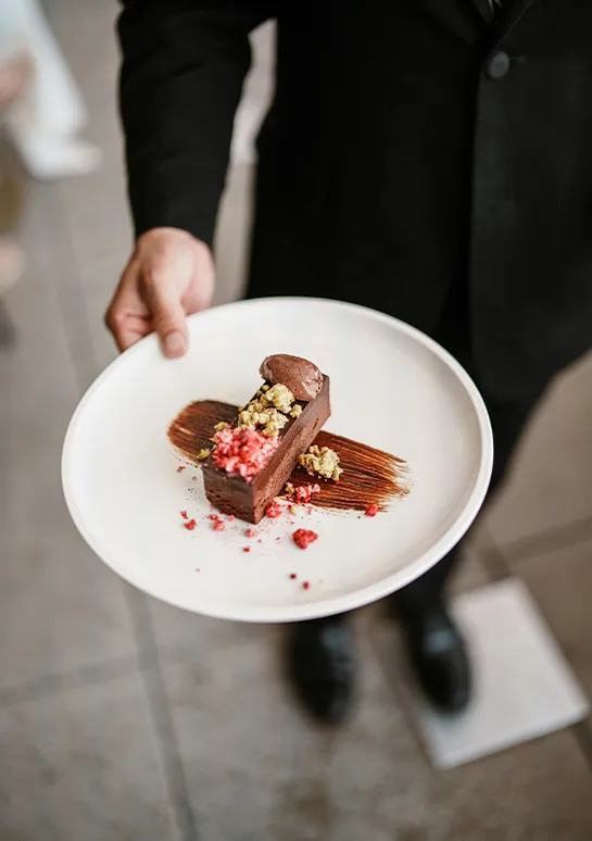 Server presenting a plated chocolate torte with pistachio and raspberry crumble