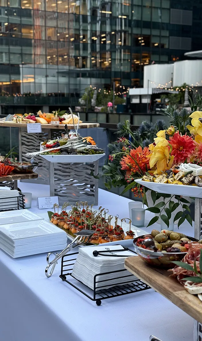 Outdoor corporate catering buffet with charcuterie, appetizers and floral arrangements beside office towers