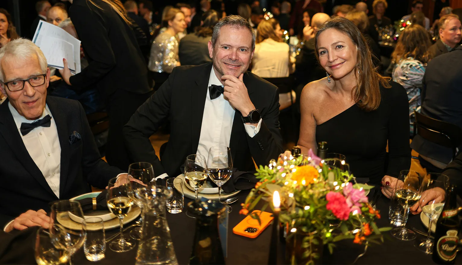 Guests in black tie seated at a candlelit gala dinner table with a floral centerpiece