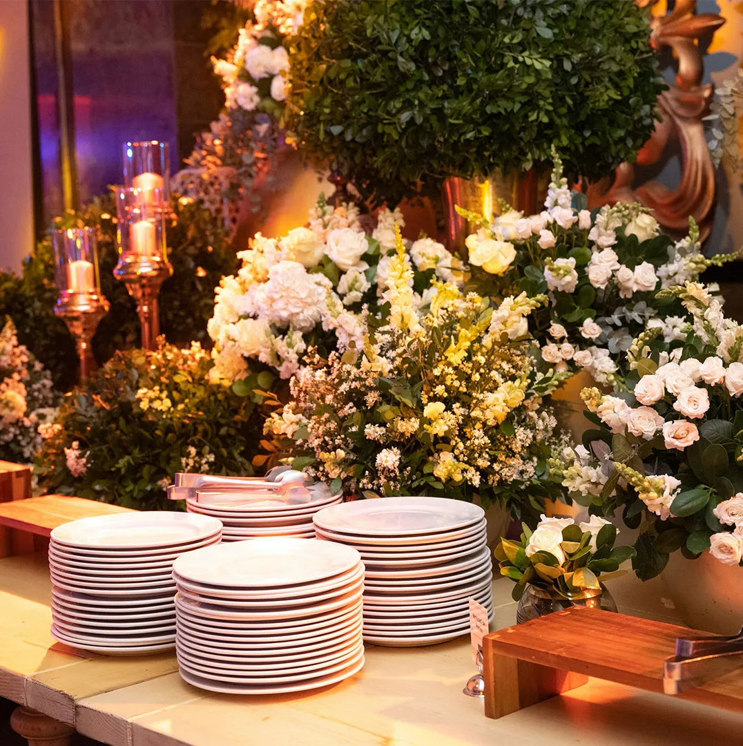 Stacked white plates on a buffet table framed by white and yellow floral arrangements and candles