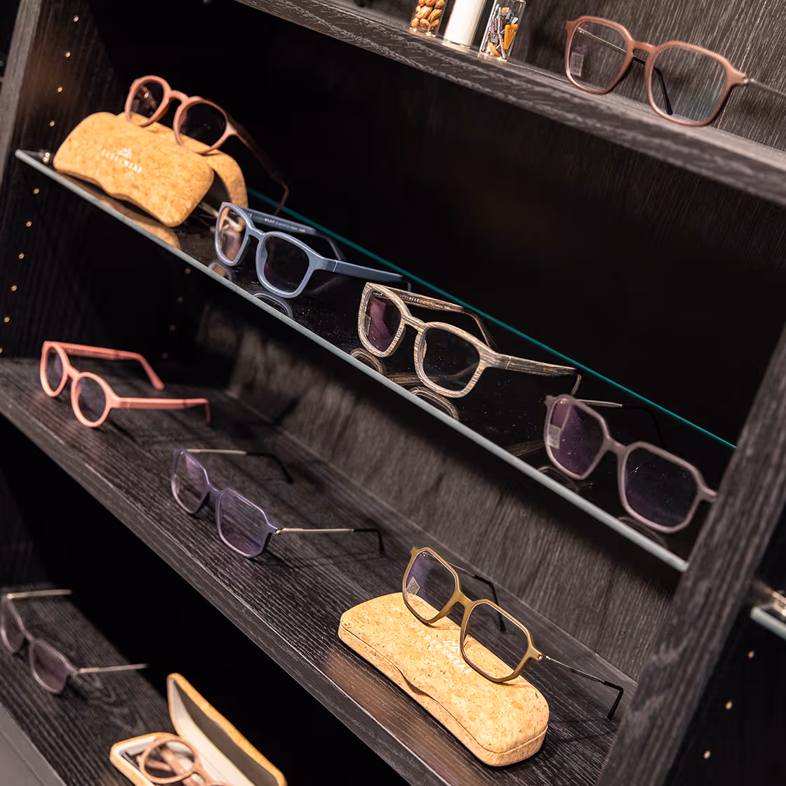 Display of various stylish eyeglass frames arranged on dark wooden shelves and a cork eyeglass case.