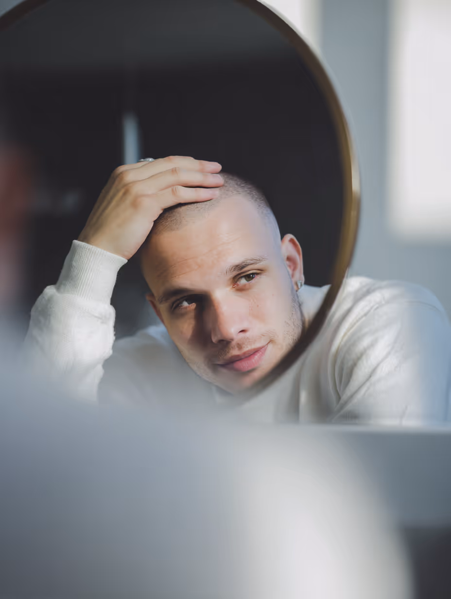 Young man with a shaved head looking thoughtfully at his reflection in a round mirror while touching his head.