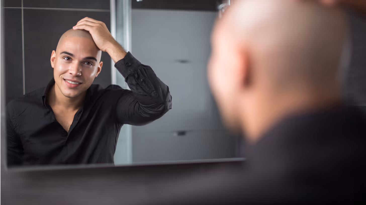 Smiling bald man in a black shirt looking at himself in a bathroom mirror with one hand on his head.