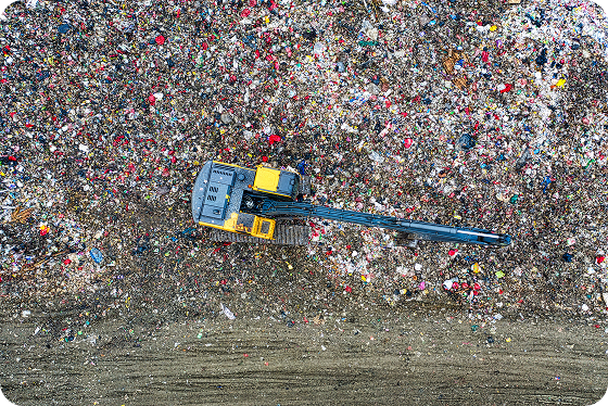 Aerial view of a large excavator with a long arm working on a vast landfill covered with scattered trash and debris.