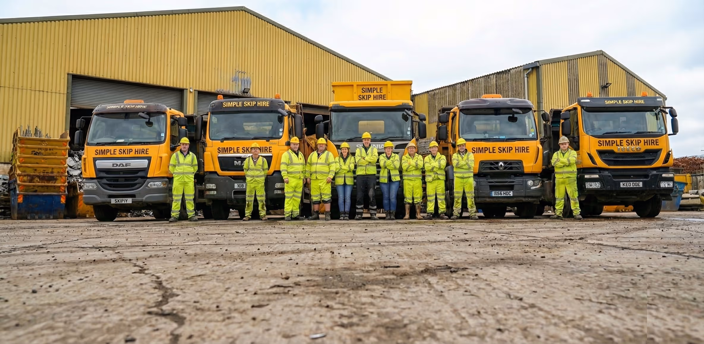 Group of workers in yellow high-visibility uniforms and hard hats standing in front of five yellow Simple Skip Hire trucks outside a warehouse.