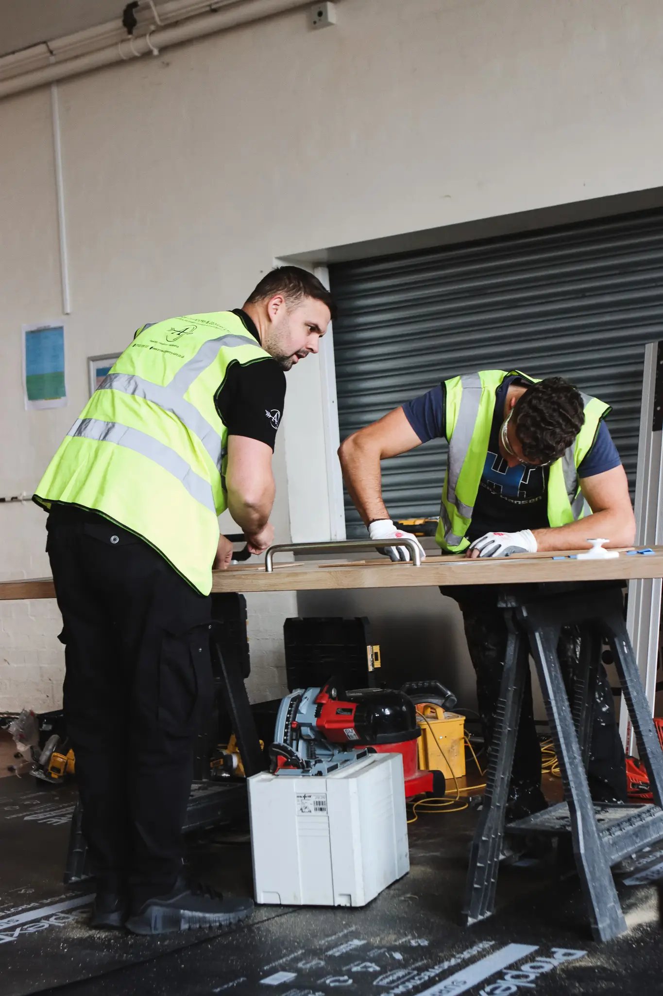 Two construction workers in yellow safety vests working on a wooden board placed on sawhorses inside a workshop.