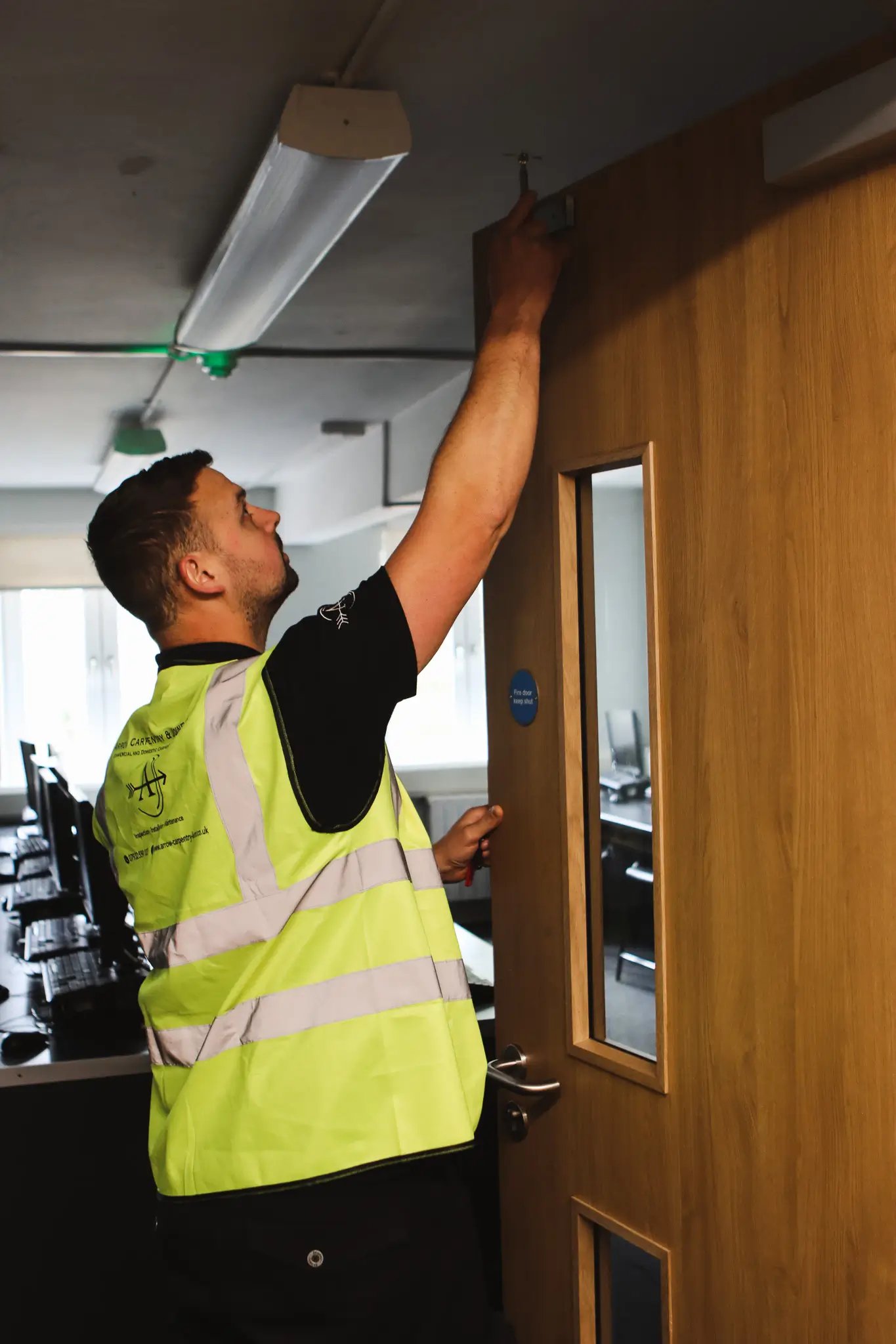 Worker in a yellow safety vest fixing a screw at the top corner of a wooden door in an office.