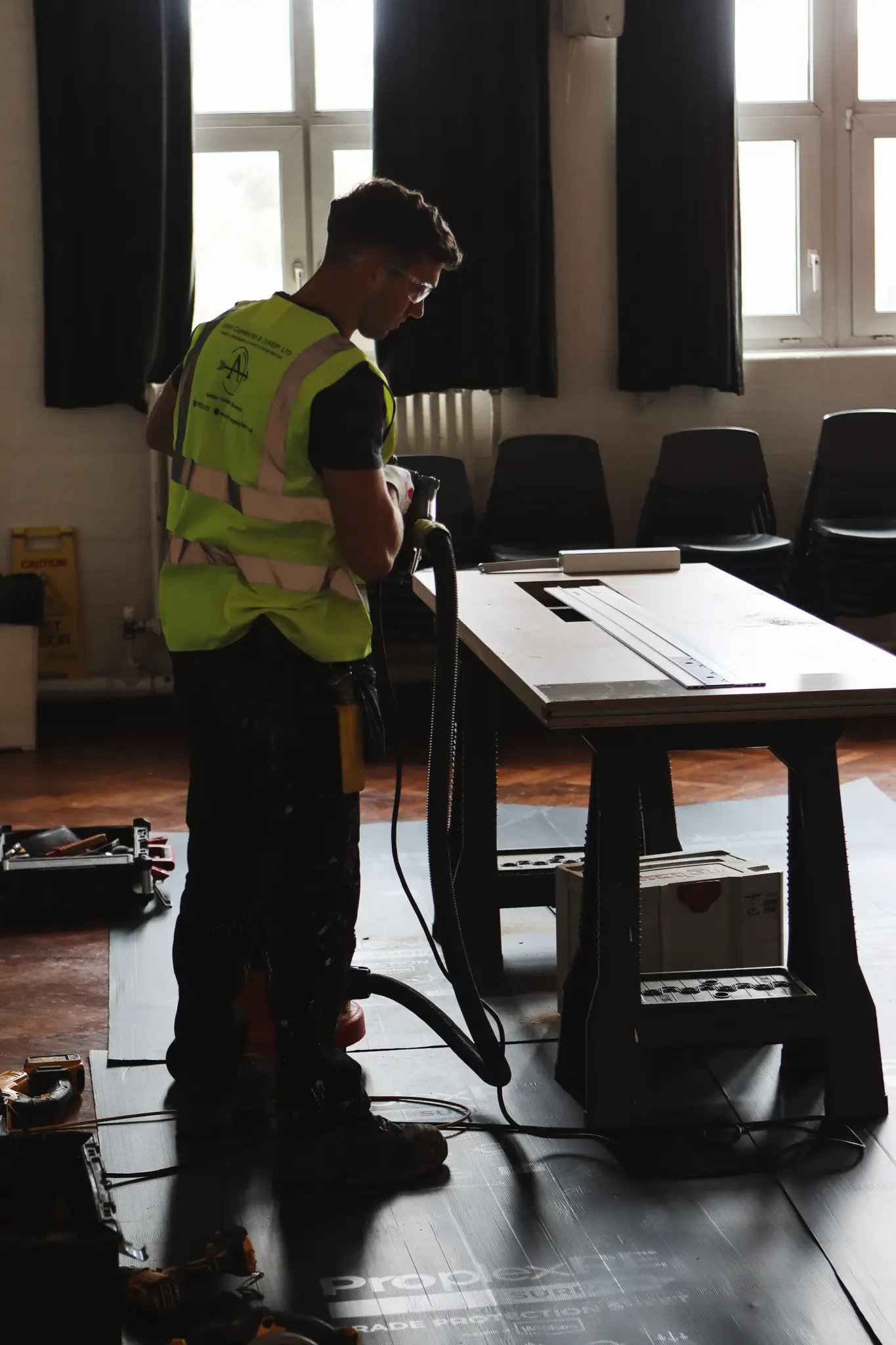 Worker in a high-visibility vest operating a power tool on a table in a workshop with tools and chairs in the background.