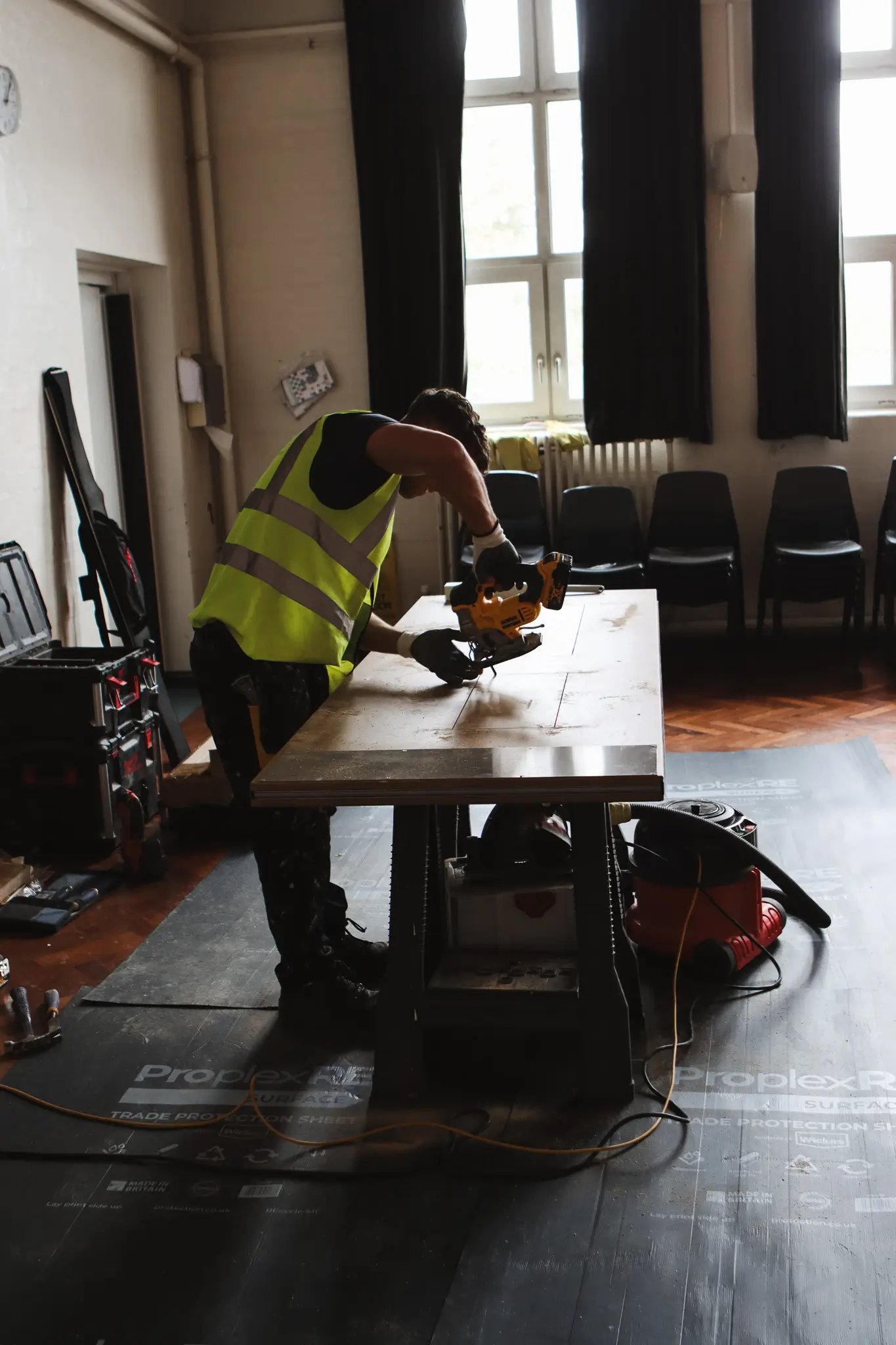 Worker in a reflective vest using a power saw to cut wood on a workbench inside a room with large windows.