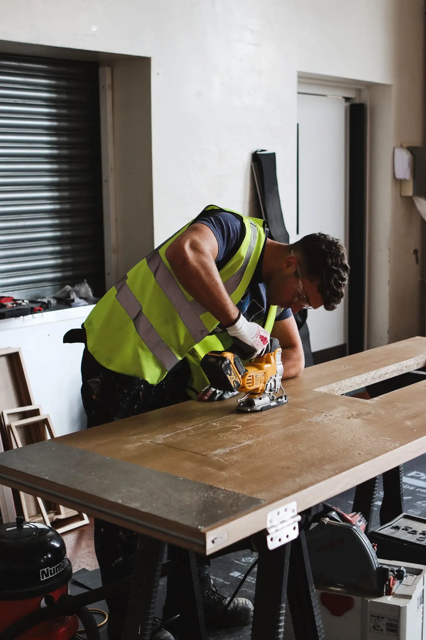 Worker in a yellow reflective vest using a power jigsaw to cut a wooden board in a workshop.