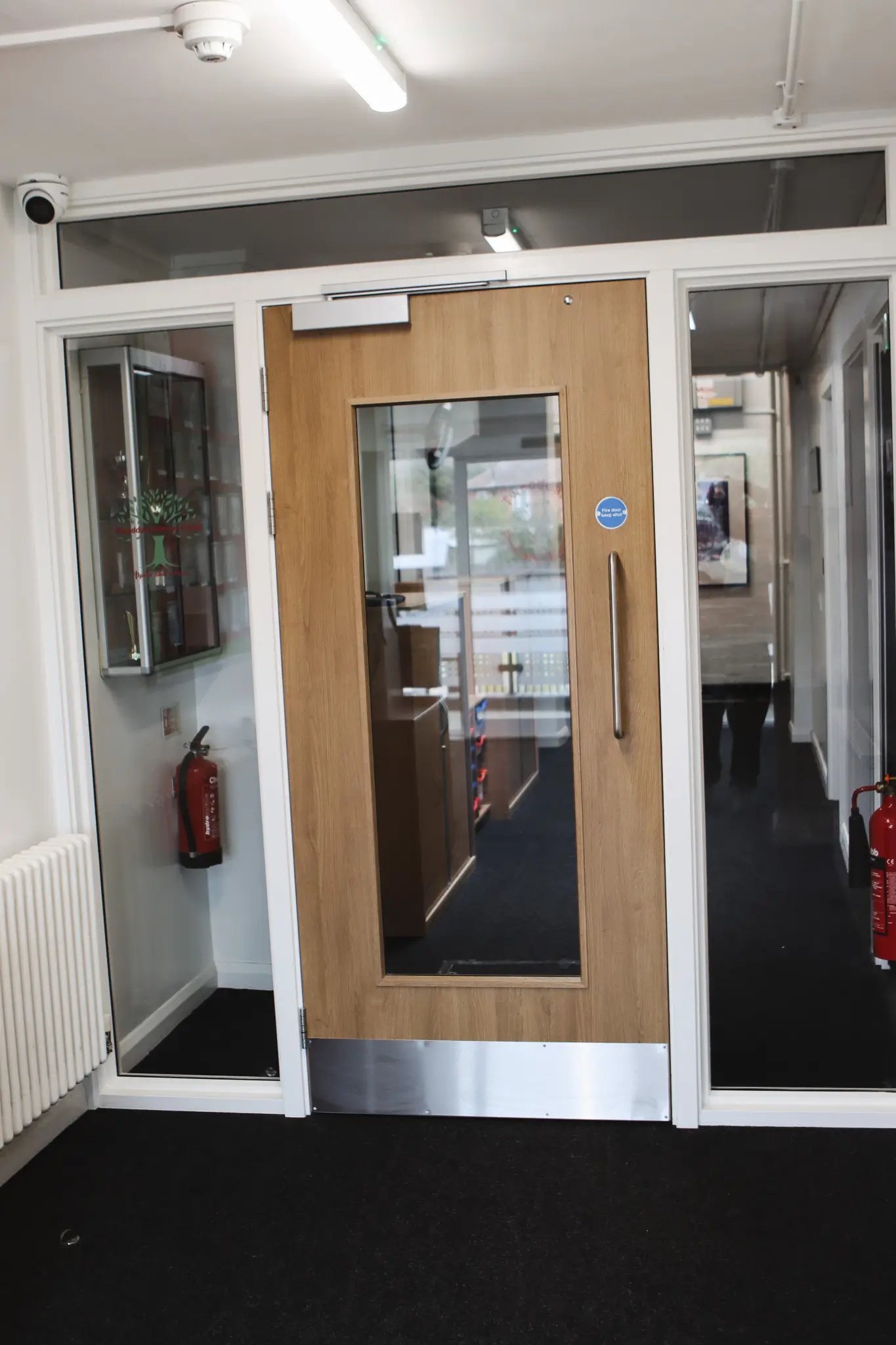 Wooden interior door with glass panel and metal kick plate in an office hallway with fire extinguishers on both sides.