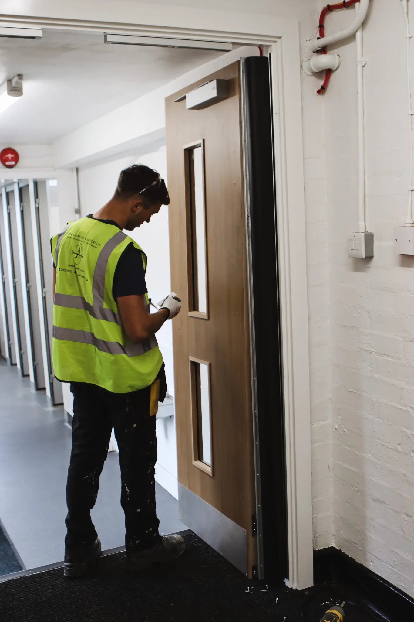 Worker in a yellow safety vest inspecting or working on a wooden door in a hallway.