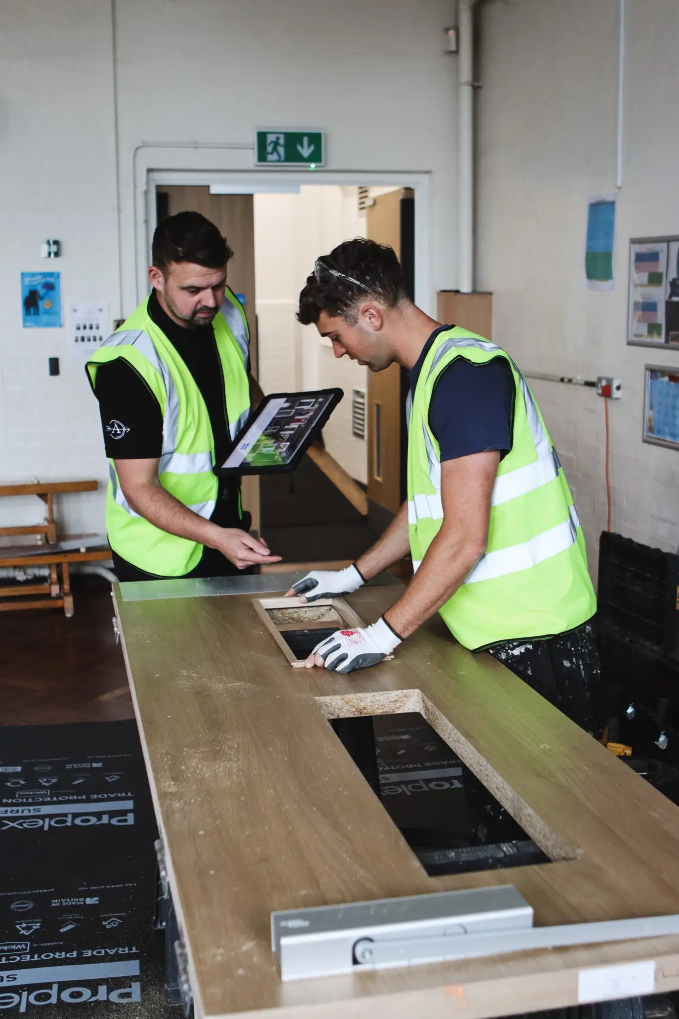 Two male workers in yellow safety vests assembling a wooden door, one holding a tablet and the other fitting a panel into a cutout.