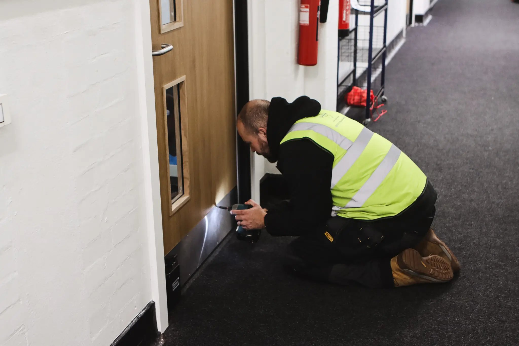 Worker in a high-visibility vest kneeling on carpeted floor using a power drill on the lower part of a wooden door inside a building.