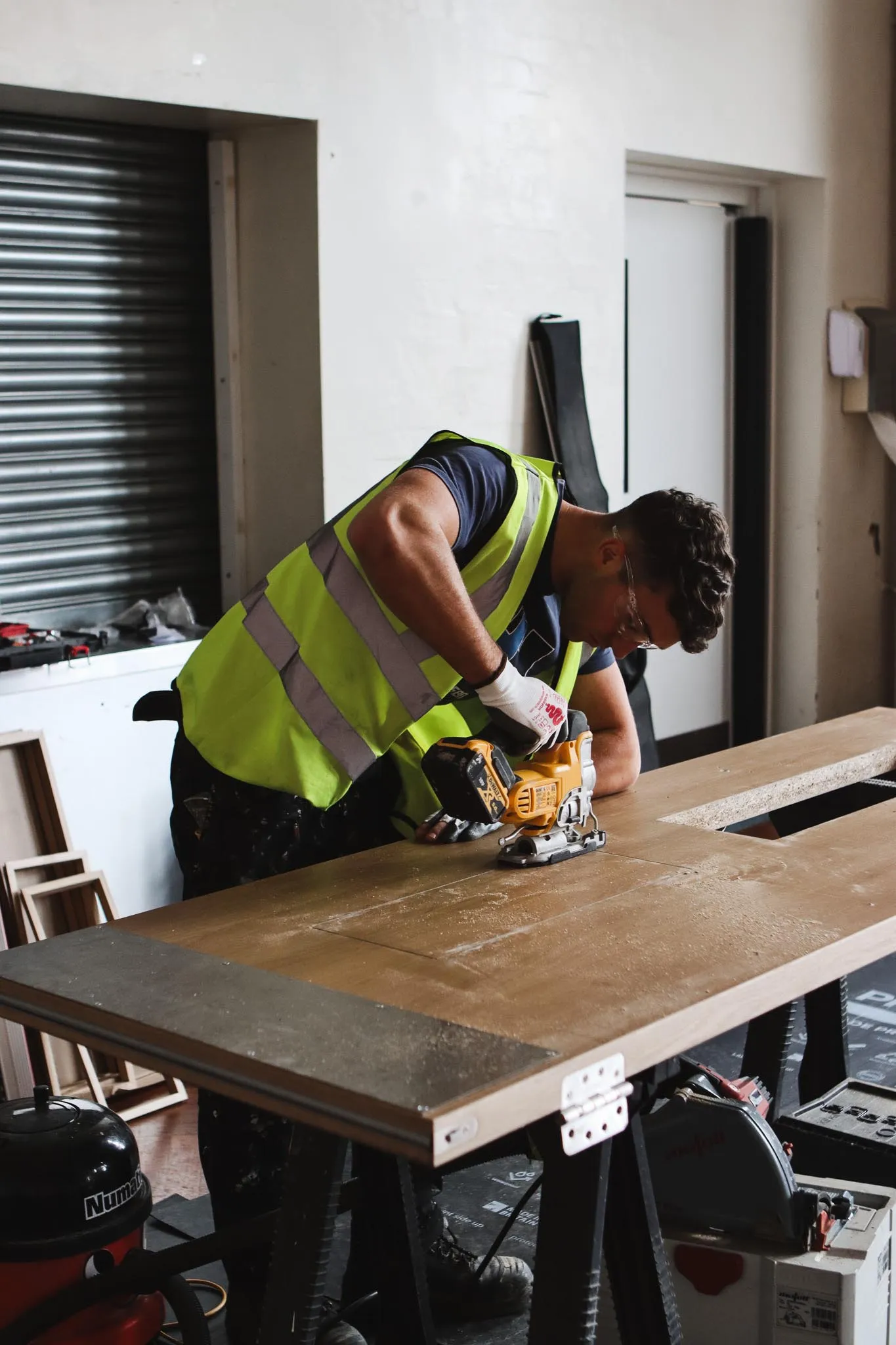 Construction worker wearing a yellow safety vest using a handheld power jigsaw to cut a piece of wood on a workbench.