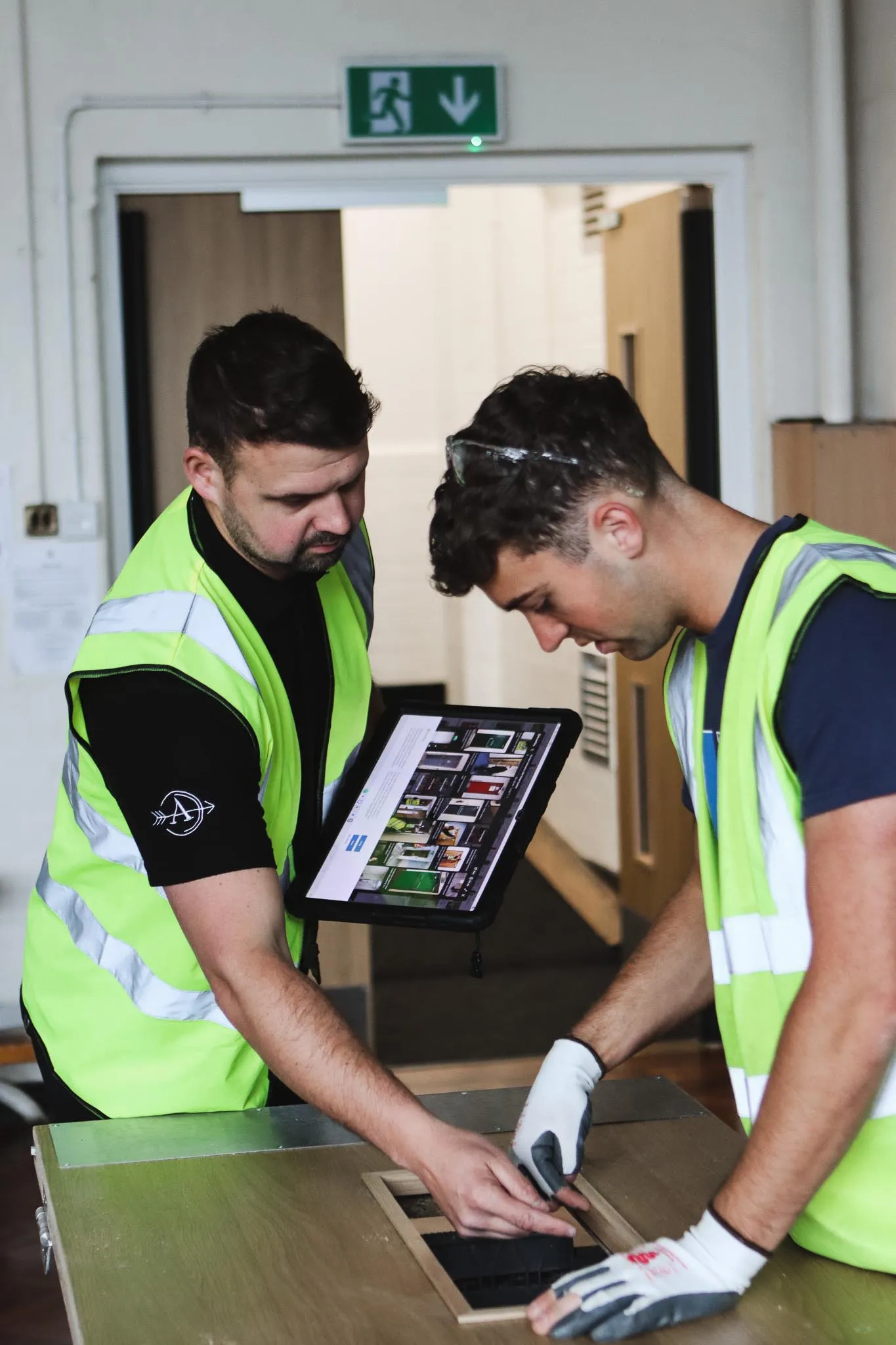Two workers in high-visibility vests examining a wooden panel, one holding a tablet displaying images.