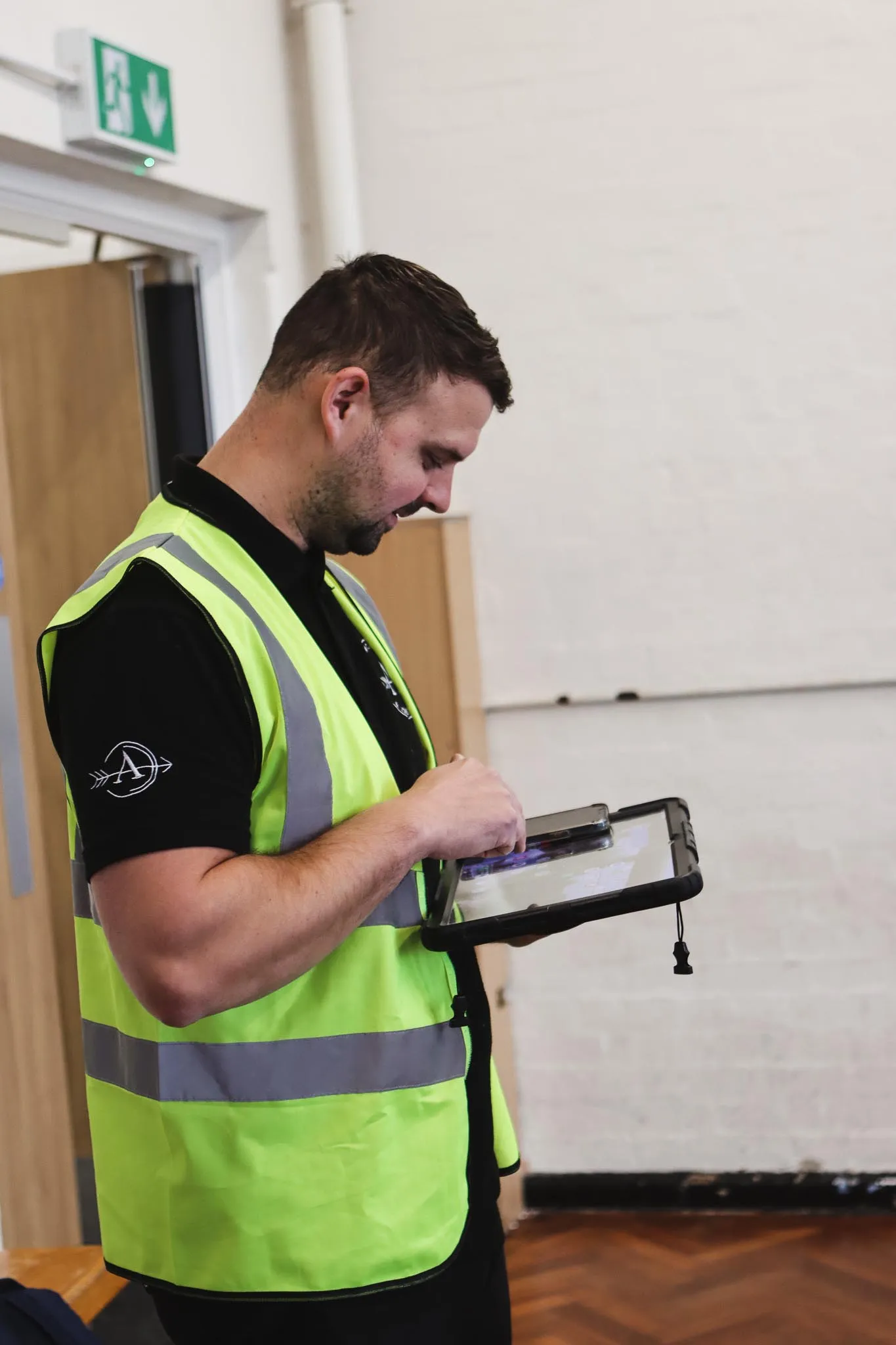Man in high-visibility vest using a tablet inside a room with wooden floor and white walls.