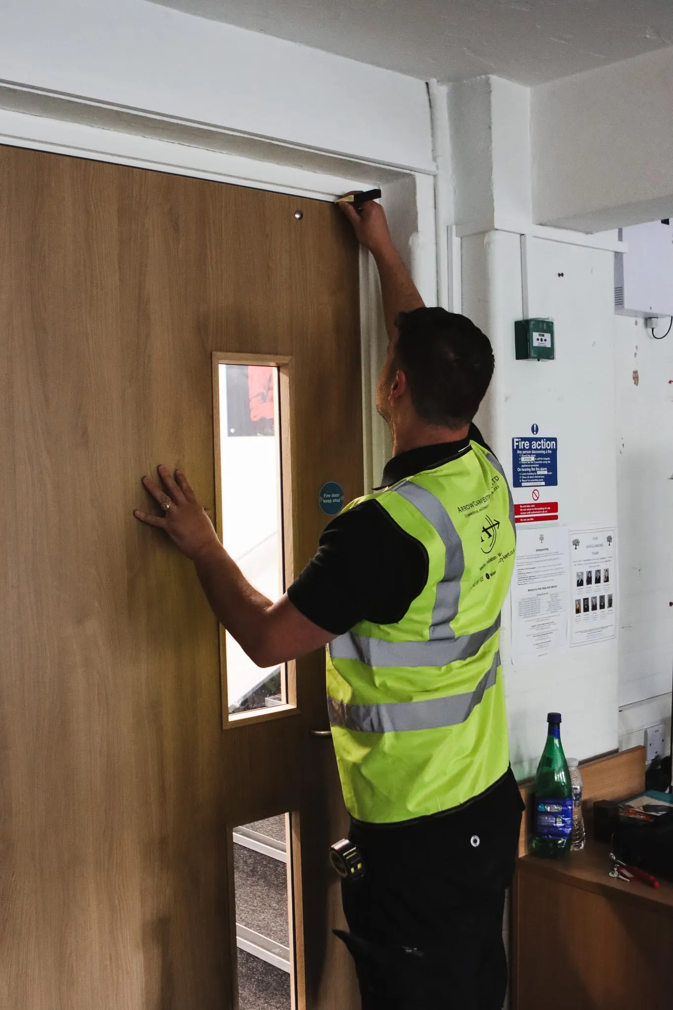 Worker in a high-visibility vest measuring or inspecting the top of a wooden door indoors.